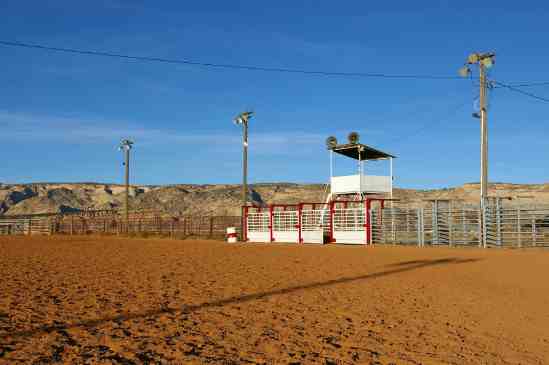 An outdoor rodeo arena with fence, chutes, and announcer tower with loudspeakers. There are desert hills behind the arena.